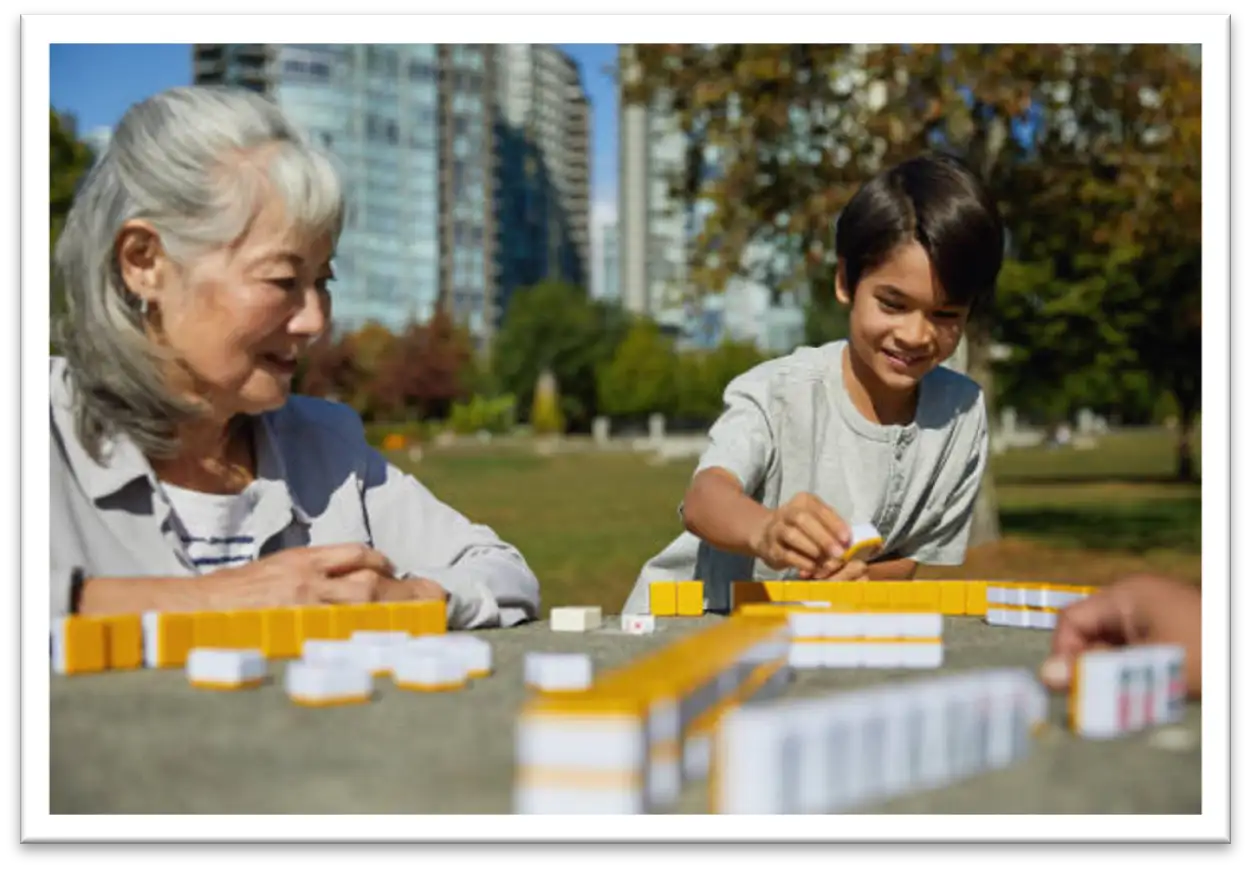 An Elderly lady and a young boy playing Mahjong outside in the park with an unseen third player.