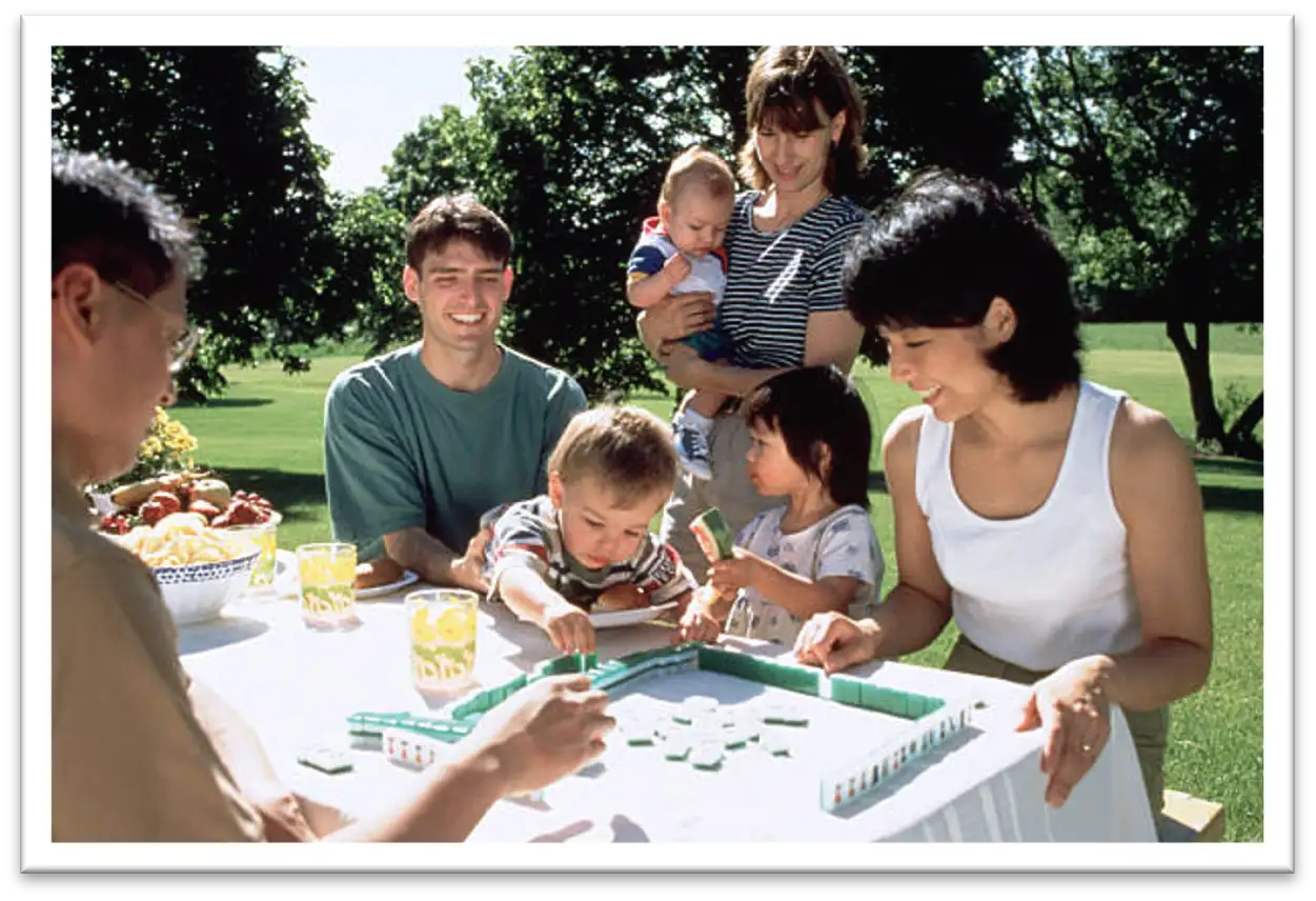 A mixed Asian/Caucasian family playing Mahjong outside at a table in a park,